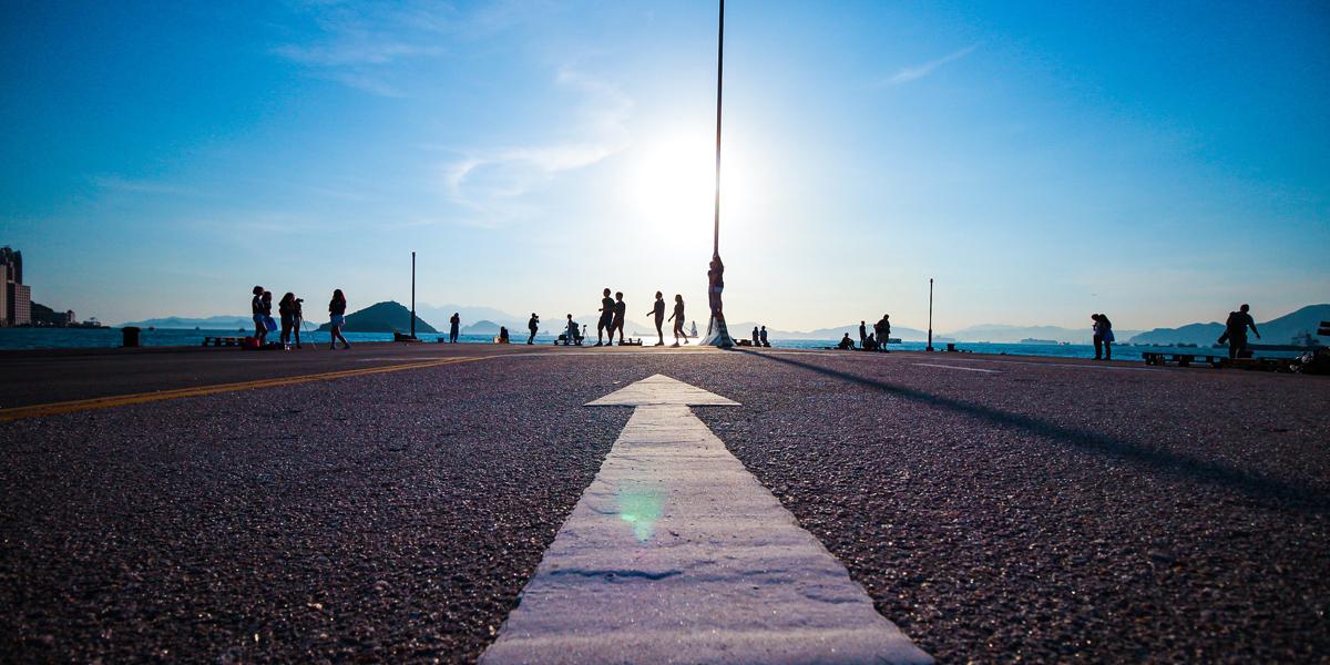 Road surface with white arrow pointing towards silhouettes of people against a bright sky.