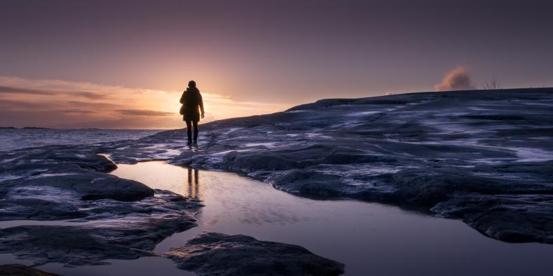Kuvassa henkilö kävelee Helsingissä meren rannassa auringonnousun aikaan.