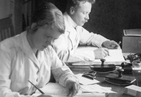 Svartvit bild från 1918, en kvinna och en man sitter vid ett skrivbord och gör anteckningar. Mannen är andelsman Otto Selenius i Oravais, kvinnan är oidentifierad.