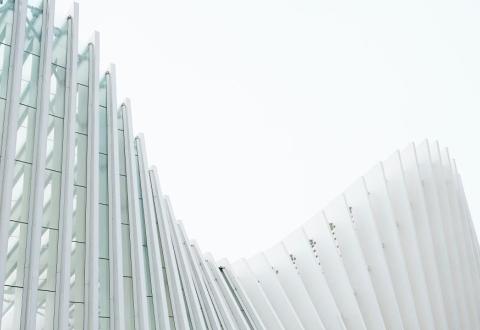 Fotografi av en välvd byggnad med raka, ljusa balkar mot vitgrå himmel..